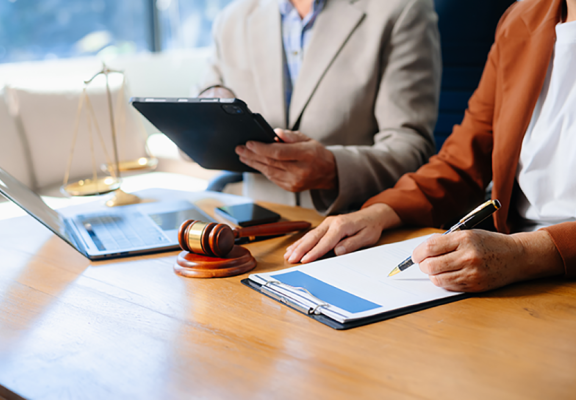 Two people in business clothing sitting at a table with a clipboard, a gavel, a laptop, a tablet, and balance scales.